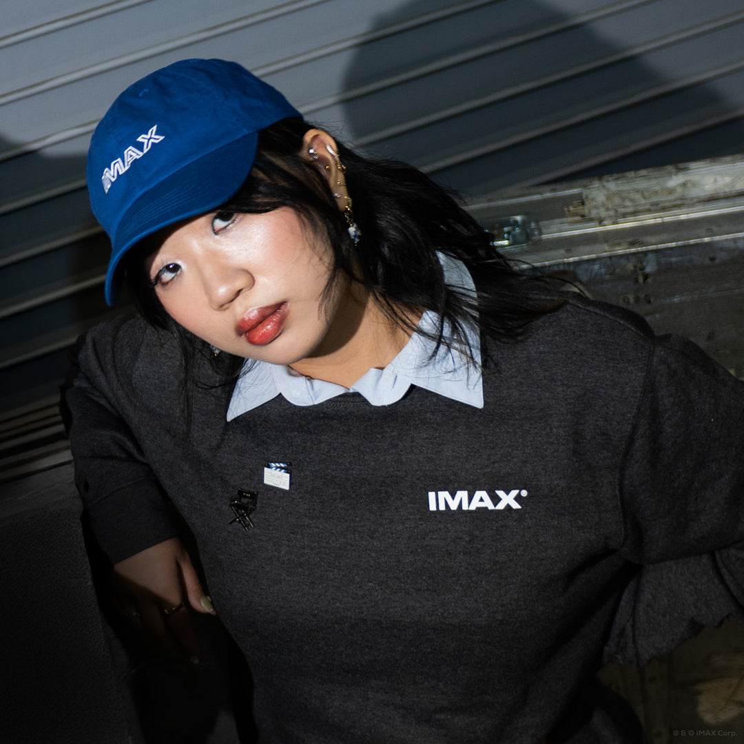 A woman posing in a loading bay wearing a blue retro-themed IMAX hat and a dark grey crewneck sweatshirt with a small white IMAX logo in front.