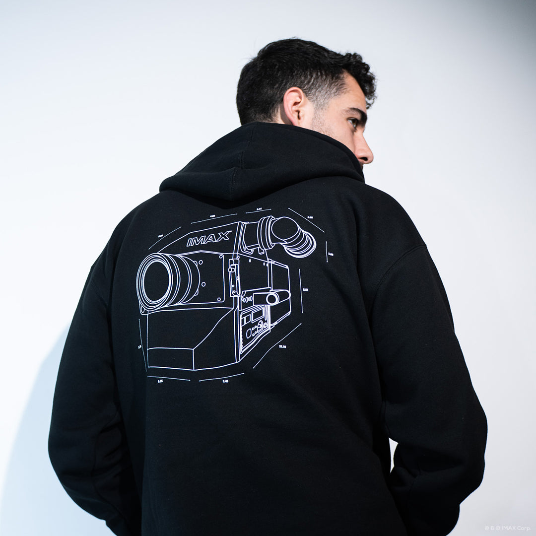 A man posing against a white wall wearing a black pullover hoodie featuring an IMAX Film Camera line art in white on the back.