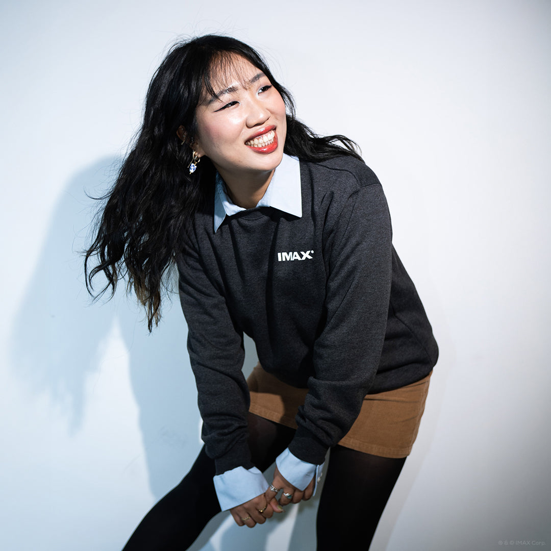 A woman posing against a white wall and smiling, wearing a dark grey crewneck sweatshirt with a small white IMAX logo over a light blue collared shirt.