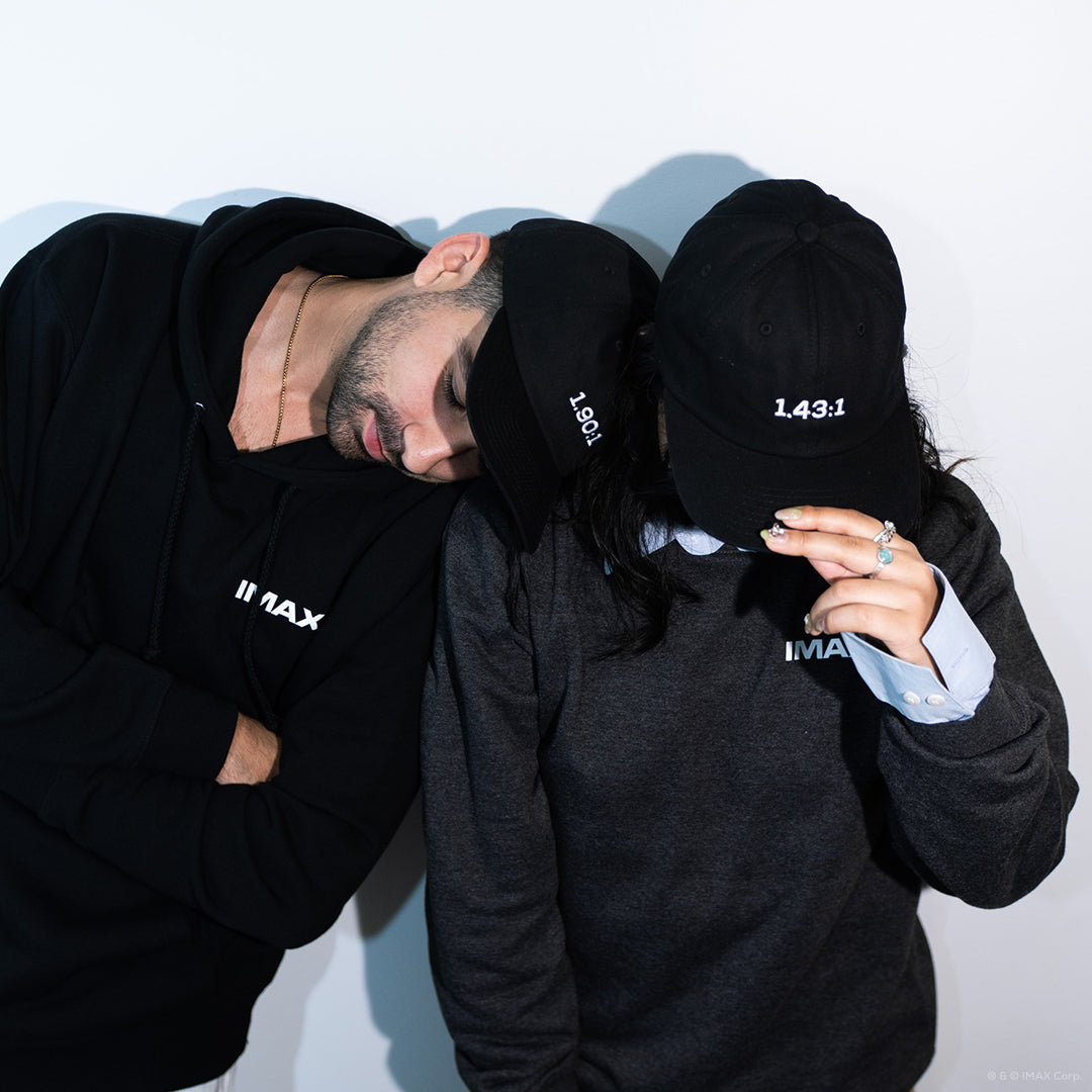 A man and woman posing against a white wall wearing various IMAX-branded apparel, including a black pullover IMAX Film Camera Hoodie, a dark-grey crewneck sweatshirt with a white IMAX logo, and black 1.90:1 and 1.43:1 Aspect Ratio Hats.