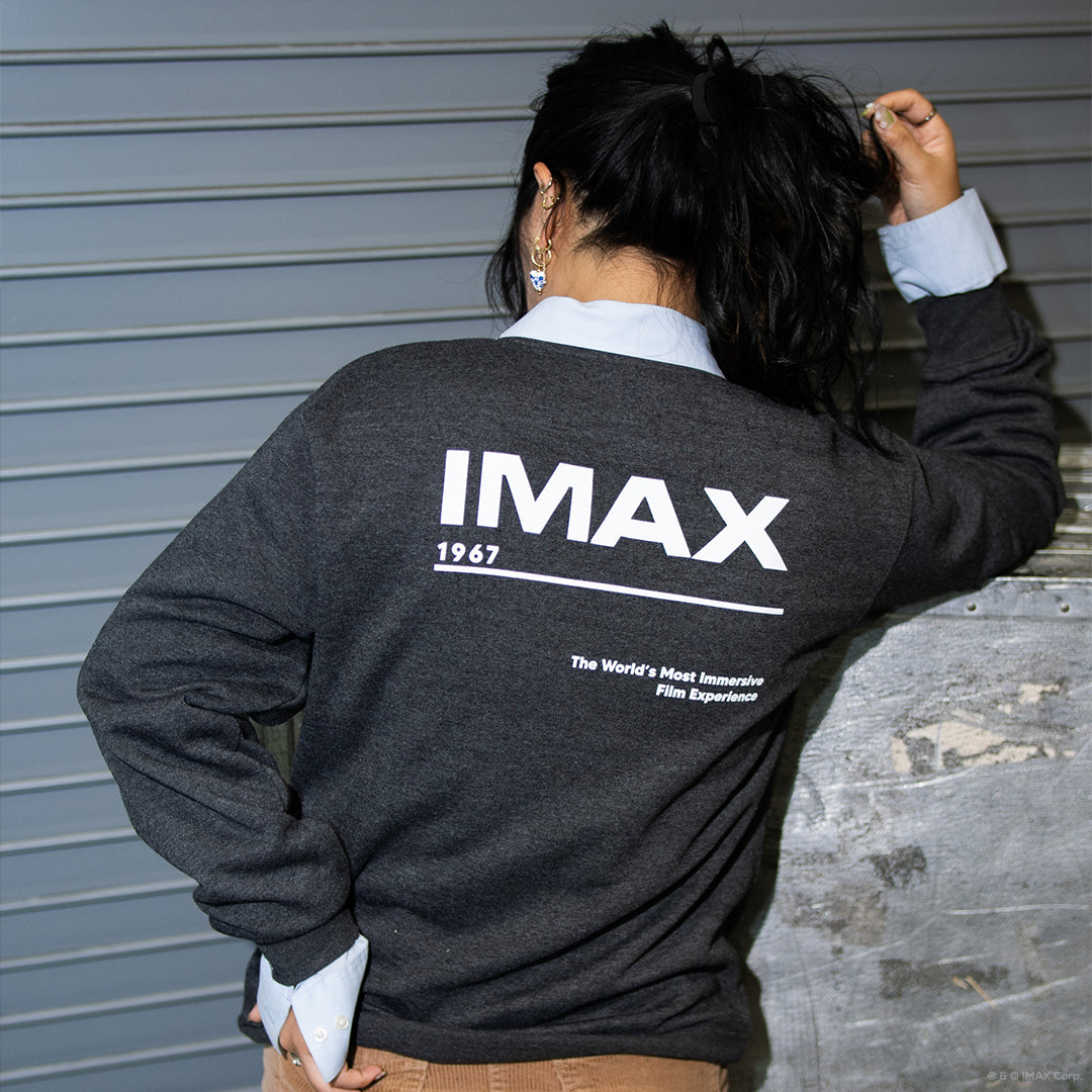 A woman posing in a loading bay, facing away from the camera and wearing a dark grey crewneck sweatshirt with a large white IMAX logo, “1967” text, a dividing line, and the phrase “The World’s Most Immersive Film Experience” on the back.
