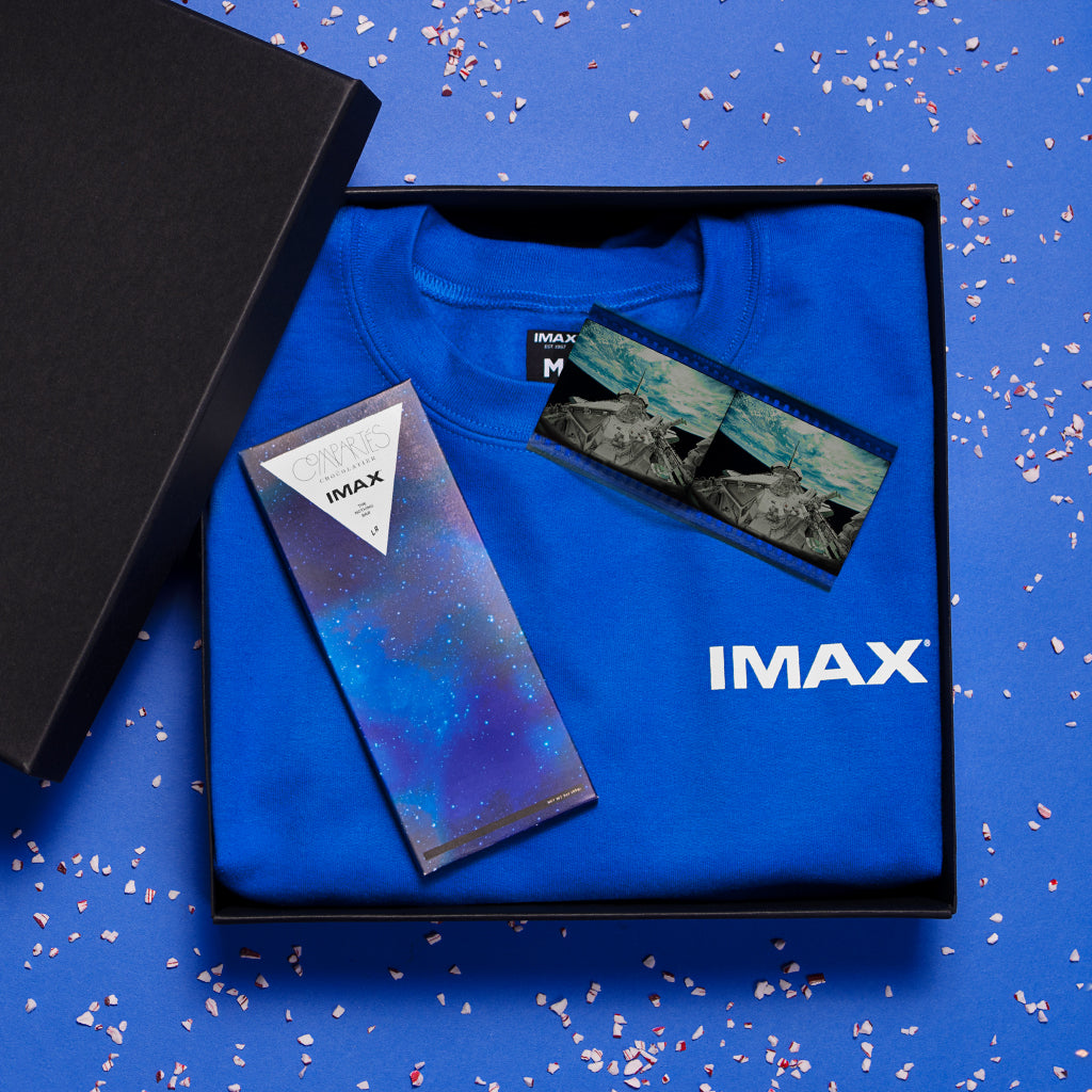 An open black gift box containing a crewneck sweatshirt from the 15/70 collection in blue, an IMAX ‘Nothing’ Bar chocolate bar from the Compartés x IMAX collection, and two frames of an IMAX 70mm print of the documentary “The Dream is Alive”.