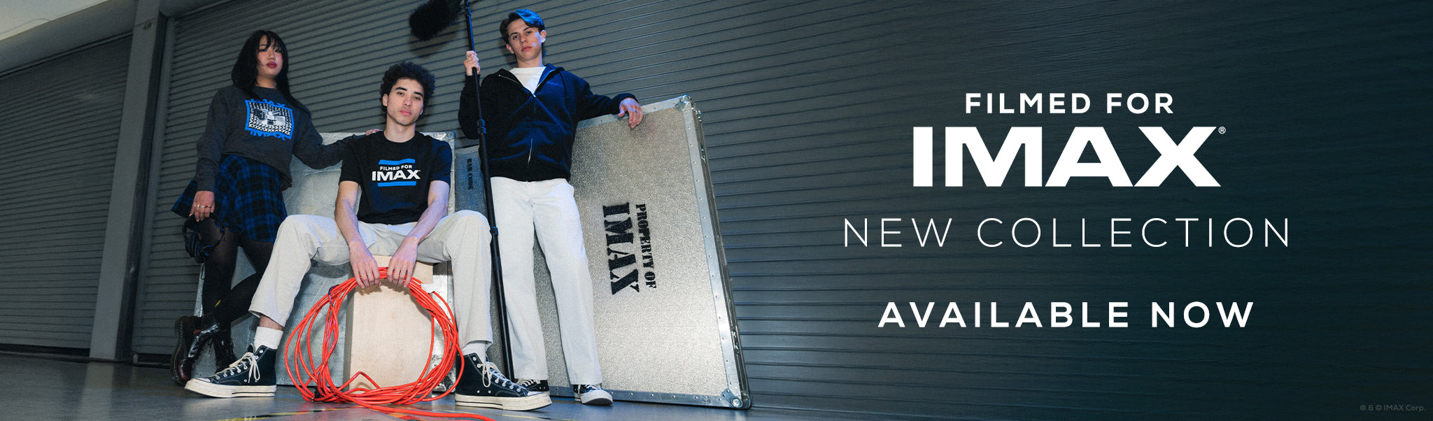 A group of two men and one woman posing with film equipment in an equipment loading bay, wearing various IMAX-branded apparel – a dark grey and blue Filmed For IMAX crewneck sweatshirt, a black and blue Filmed For IMAX T-shirt, and an all-black IMAX-branded hoodie.  