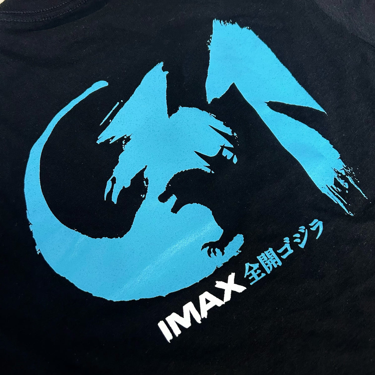Close-up of a black T-shirt featuring a large light blue godzilla graphic and white Japanese text reading 'IMAX 全開ゴジラ'.