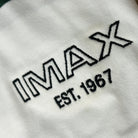 Close of up the embroidery of a Green and white striped rugby style long sleeve polo with a white collar. Green embroidered IMAX est 1967 small on the left chest. They are also wearing a matching green and cream hat with IMAX logo on the front. 