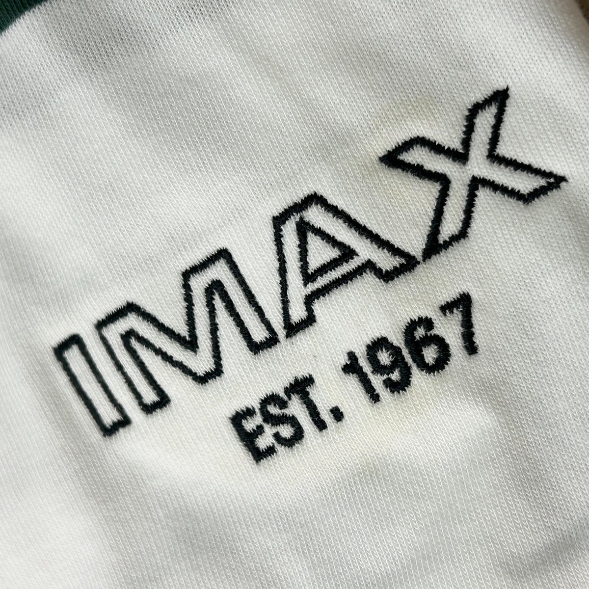 Close of up the embroidery of a Green and white striped rugby style long sleeve polo with a white collar. Green embroidered IMAX est 1967 small on the left chest. They are also wearing a matching green and cream hat with IMAX logo on the front. 