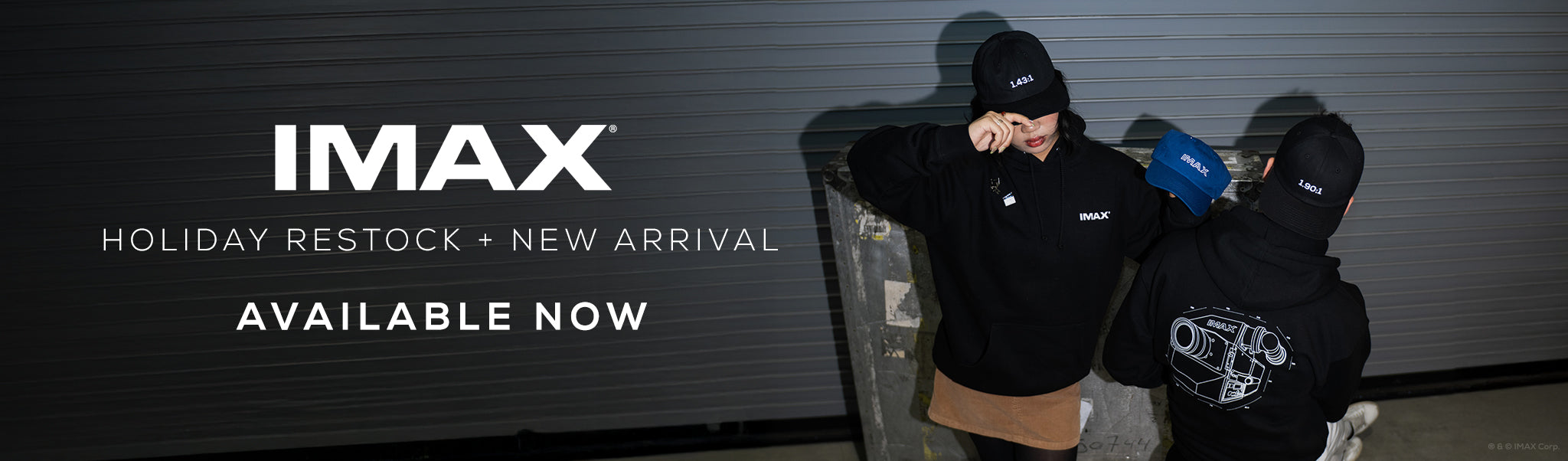 A banner featuring a man and woman posing in a loading bay wearing various IMAX-branded apparel, including a black pullover IMAX Film Camera Hoodie, black 1.90:1 and 1.43:1 Aspect Ratio Hats, and a blue retro-themed IMAX Hat. Banner copy reads IMAX Holiday Restock + New Arrival Available Now” in white text.