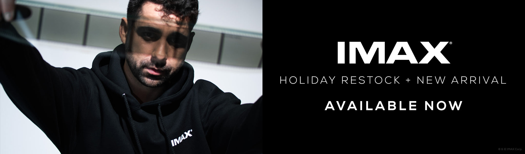 A banner with a man posing against a white wall, holding an IMAX 70mm Film filmstrip and wearing a black pullover hoodie with a white IMAX logo in front. Banner copy reads IMAX Holiday Restock + New Arrival Available Now” in white text. 