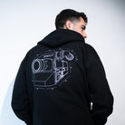 A man posing against a white wall wearing a black pullover hoodie featuring an IMAX Film Camera line art in white on the back. 