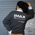 A woman posing in a loading bay, facing away from the camera and wearing a dark grey crewneck sweatshirt with a large white IMAX logo, “1967” text, a dividing line, and the phrase “The World’s Most Immersive Film Experience” on the back.