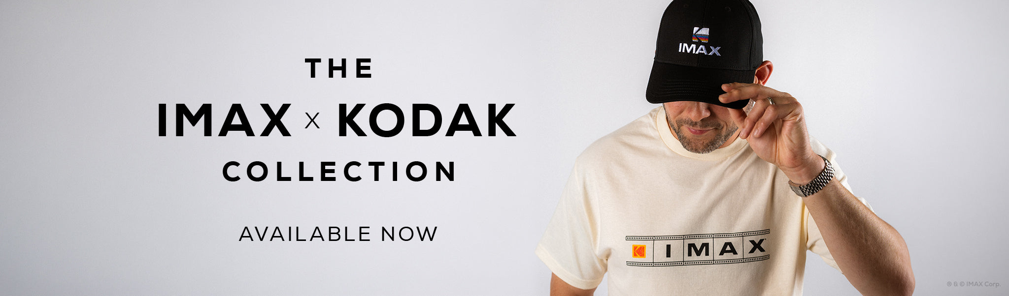 Person wearing a Ccream IMAX short-sleeve T-shirt with horizontal filmstrip graphic, orange Kodak logo, and black IMAX logo across the chest. They have their hand on the brim of a black hat that has the Kodak logo and IMAX written under the logo. Text reads "The IMAX x KODAK COLLECTION AVAILABLE NOW"