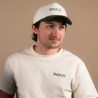 Person wearing a cream shirt with a small green imax embroidered logo on the left chest and a matching cream hat with a green bill and green embroidered IMAX logo on the front