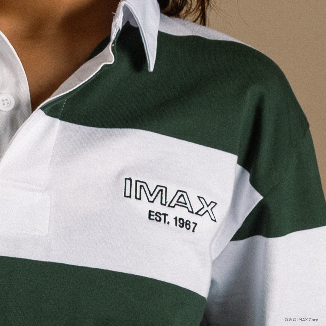 Close of of the left chest of a Green and white striped rugby style long sleeve polo with a white collar. Green embroidered IMAX est 1967 small on the left chest. They are also wearing a matching green and cream hat with IMAX logo on the front. 